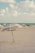 A sandy beach setting with a large umbrella labeled 'Esteban's Place' positioned prominently in the foreground. Several people are seen relaxing on the beach in the background near the shoreline. The sky is partly cloudy, and the ocean water is visible in the distance.
