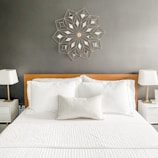 The image features a well-kept bedroom with a neatly arranged bed and two white pillows, set against a gray wall. Above the bed is a decorative wall art in a geometric pattern. On each side of the bed, there are white nightstands with modern lamps featuring white lampshades.