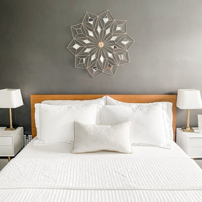 The image features a well-kept bedroom with a neatly arranged bed and two white pillows, set against a gray wall. Above the bed is a decorative wall art in a geometric pattern. On each side of the bed, there are white nightstands with modern lamps featuring white lampshades.