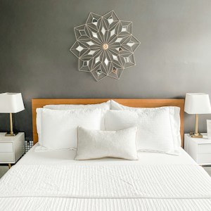 The image features a well-kept bedroom with a neatly arranged bed and two white pillows, set against a gray wall. Above the bed is a decorative wall art in a geometric pattern. On each side of the bed, there are white nightstands with modern lamps featuring white lampshades.
