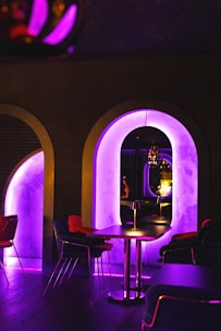 A dimly lit room features modern furniture including tables and chairs. The focal point is an archway illuminated with a neon purple light, reflecting onto another archway and creating a futuristic ambiance. Hanging light fixtures and a cozy seating arrangement complete the scene.