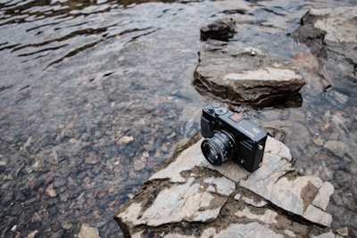 A digital camera sits on a large rock near the edge of a shallow, rocky body of water. The camera appears to be a compact model, with a lens facing forward. The water is clear, revealing an assortment of small stones and pebbles underneath. The setting has a natural, outdoor feel with rock formations surrounding the scene.