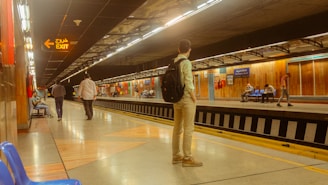 A subway station platform with several people waiting. One person stands with a backpack, while others sit on benches or walk along the platform. There are overhead lights illuminating the path, with an exit sign in both English and Arabic. The walls are mostly wooden panels, adding a warm tone to the scene.