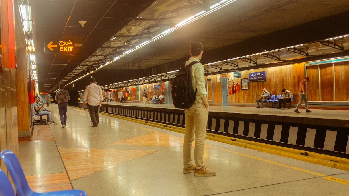 A subway station platform with several people waiting. One person stands with a backpack, while others sit on benches or walk along the platform. There are overhead lights illuminating the path, with an exit sign in both English and Arabic. The walls are mostly wooden panels, adding a warm tone to the scene.