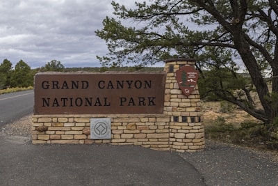 The image features a large stone entrance sign for Grand Canyon National Park, surrounded by trees and a paved road leading into the park. The sign includes a brown rectangular plaque with the park's name in bold letters, and a circular emblem underneath recognizing the site's world heritage status. A tree with lush green foliage adds a natural element to the scene.