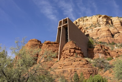 A modern building with a tall, vertical structure is integrated into a rocky, desert landscape. The building is made of concrete and features a triangular cut at the top. It is surrounded by red rock formations and sparse desert vegetation.