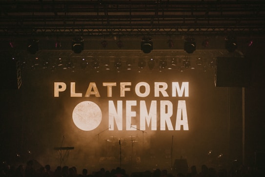 A dimly lit stage with the words 'PLATFORM NEMRA' displayed in large, bold letters across the background screen. Musical instruments such as a keyboard, drums, and microphones are visible on the stage. Stage lights hang from above, contributing to the atmospheric lighting.