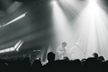 A live music performance with musicians playing on stage, surrounded by dramatic lighting and a crowd in the foreground. The atmosphere is dynamic, with beams of light cutting through hazy air, creating an intense and immersive experience.