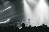 A live music performance with musicians playing on stage, surrounded by dramatic lighting and a crowd in the foreground. The atmosphere is dynamic, with beams of light cutting through hazy air, creating an intense and immersive experience.