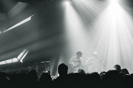 A live music performance with musicians playing on stage, surrounded by dramatic lighting and a crowd in the foreground. The atmosphere is dynamic, with beams of light cutting through hazy air, creating an intense and immersive experience.