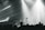 A live music performance with musicians playing on stage, surrounded by dramatic lighting and a crowd in the foreground. The atmosphere is dynamic, with beams of light cutting through hazy air, creating an intense and immersive experience.