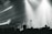 A live music performance with musicians playing on stage, surrounded by dramatic lighting and a crowd in the foreground. The atmosphere is dynamic, with beams of light cutting through hazy air, creating an intense and immersive experience.