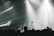 A live music performance with musicians playing on stage, surrounded by dramatic lighting and a crowd in the foreground. The atmosphere is dynamic, with beams of light cutting through hazy air, creating an intense and immersive experience.