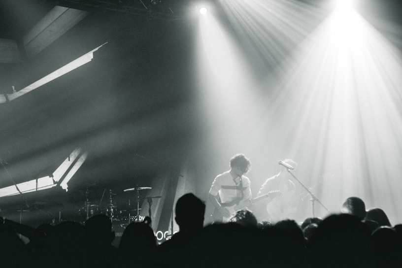 A live music performance with musicians playing on stage, surrounded by dramatic lighting and a crowd in the foreground. The atmosphere is dynamic, with beams of light cutting through hazy air, creating an intense and immersive experience.