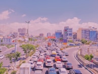 A busy urban traffic scene with numerous cars lined up on multiple lanes in the foreground. Modern buildings and skyscrapers make up the background, with mountains visible in the distance. The sky is clear with a few clouds, and there are trees and some green spaces along the road.