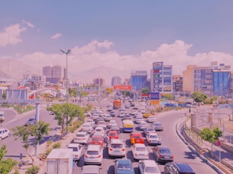A busy urban traffic scene with numerous cars lined up on multiple lanes in the foreground. Modern buildings and skyscrapers make up the background, with mountains visible in the distance. The sky is clear with a few clouds, and there are trees and some green spaces along the road.