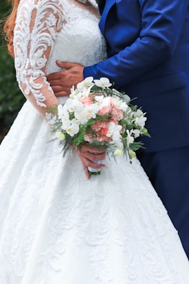 A bride in an ornate white wedding dress with lace detailing is holding a bouquet of pink and white flowers with green foliage. She is being embraced by a person in a blue suit, creating an intimate and romantic atmosphere.