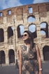 The image features a woman wearing sunglasses and a patterned dress standing in front of an ancient stone structure with large arches. The sky is partly cloudy.