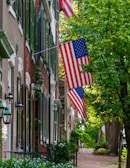 A quaint, tree-lined street features a series of historic brick townhouses adorned with American flags. Green shutters and black lanterns add charm, while lush foliage and cobblestone pathways create a serene urban setting.