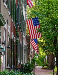 A quaint, tree-lined street features a series of historic brick townhouses adorned with American flags. Green shutters and black lanterns add charm, while lush foliage and cobblestone pathways create a serene urban setting.