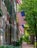 A quaint, tree-lined street features a series of historic brick townhouses adorned with American flags. Green shutters and black lanterns add charm, while lush foliage and cobblestone pathways create a serene urban setting.