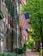 A quaint, tree-lined street features a series of historic brick townhouses adorned with American flags. Green shutters and black lanterns add charm, while lush foliage and cobblestone pathways create a serene urban setting.