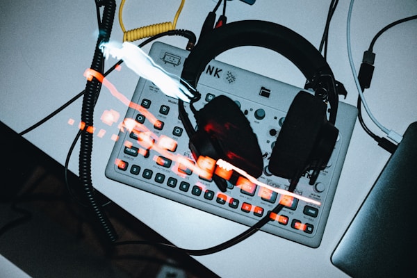 A pair of black headphones rests on top of a compact music synthesizer or drum machine, which has various buttons and dials. Cables are plugged into the device, one of which is yellow, and the environment seems to include a laptop partially visible to the side. Red and white light streaks create a dynamic effect across the image.