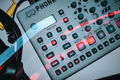 A close-up view of an electronic music production device with multiple buttons and knobs. The device has various labeled buttons such as 'Kick', 'Snare', 'Metal', 'Perc', and 'Tone'. Several knobs are arranged in rows, and there are colorful light streaks across the image. To the left, coiled cables and part of a headphone are visible.