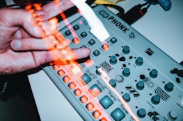 A close-up view of a light-colored electronic music device with numerous buttons, knobs, and illuminated controls. A hand, blurred with motion, is interacting with the device, creating an impression of activity and engagement in music creation.