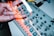 A close-up view of a light-colored electronic music device with numerous buttons, knobs, and illuminated controls. A hand, blurred with motion, is interacting with the device, creating an impression of activity and engagement in music creation.
