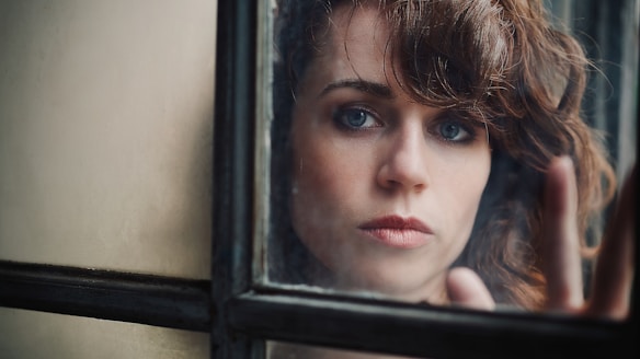 A woman with curly brown hair and blue eyes is staring intently through a window. Her expression conveys depth and introspection, with her hand gently touching the glass pane. The background consists of an out-of-focus interior with a muted color palette.