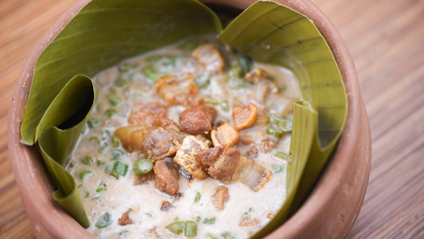 A rustic terracotta bowl lined with banana leaves contains a creamy stew with chunks of meat and green vegetables.