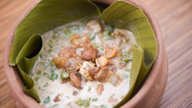 A rustic terracotta bowl lined with banana leaves contains a creamy stew with chunks of meat and green vegetables.