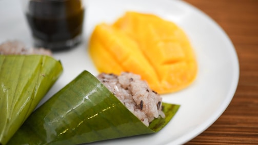 A white plate holds a serving of sticky rice wrapped in a green banana leaf, alongside sliced ripe mango. In the background, there is a small glass of a dark liquid, possibly soy sauce or a similar condiment. The wooden table surface enhances the presentation, creating a traditional, rustic vibe.