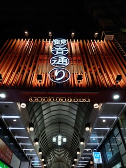 A brightly illuminated entrance to a shopping arcade or street with a curved, clear roof structure. The sign features Japanese characters in neon lights and an English translation reading 'KANNONJI DORI'. The overall setting is urban with a combination of modern architectural elements and vibrant lighting.