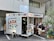 A small food truck labeled 'La Cabina' is parked on the side of a street. It offers various foods like tacos and drinks such as cold beer. There are people sitting on stools and benches nearby, enjoying their meals. Some potted plants add a touch of greenery to the scene. The atmosphere looks casual and relaxed.