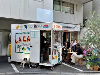 A small food truck labeled 'La Cabina' is parked on the side of a street. It offers various foods like tacos and drinks such as cold beer. There are people sitting on stools and benches nearby, enjoying their meals. Some potted plants add a touch of greenery to the scene. The atmosphere looks casual and relaxed.