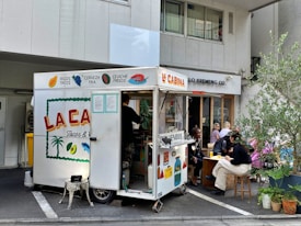 A small food truck labeled 'La Cabina' is parked on the side of a street. It offers various foods like tacos and drinks such as cold beer. There are people sitting on stools and benches nearby, enjoying their meals. Some potted plants add a touch of greenery to the scene. The atmosphere looks casual and relaxed.