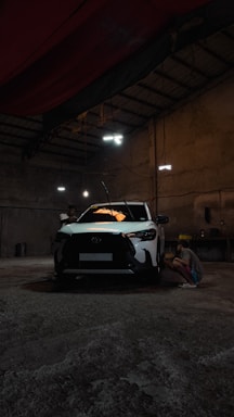 A white car is parked inside a dimly lit garage. A person is crouched near the front passenger side, possibly inspecting or working on the vehicle. The setting appears industrial with concrete floors, high ceilings, and minimal light fixtures creating a shadowy atmosphere.