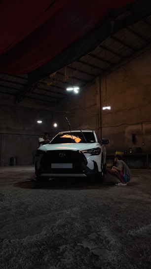 A white car is parked inside a dimly lit garage. A person is crouched near the front passenger side, possibly inspecting or working on the vehicle. The setting appears industrial with concrete floors, high ceilings, and minimal light fixtures creating a shadowy atmosphere.