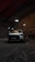 A white car is parked inside a dimly lit garage. A person is crouched near the front passenger side, possibly inspecting or working on the vehicle. The setting appears industrial with concrete floors, high ceilings, and minimal light fixtures creating a shadowy atmosphere.