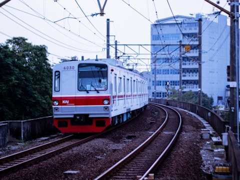 A commuter train travels along a curved railway track with overhead wires. The train is predominantly white with red accents and '6030' marked on its front. The background includes urban buildings and greenery, with a multi-story structure directly behind the tracks.