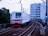 A commuter train travels along a curved railway track with overhead wires. The train is predominantly white with red accents and '6030' marked on its front. The background includes urban buildings and greenery, with a multi-story structure directly behind the tracks.