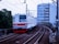 A commuter train travels along a curved railway track with overhead wires. The train is predominantly white with red accents and '6030' marked on its front. The background includes urban buildings and greenery, with a multi-story structure directly behind the tracks.