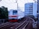 A commuter train travels along a curved railway track with overhead wires. The train is predominantly white with red accents and '6030' marked on its front. The background includes urban buildings and greenery, with a multi-story structure directly behind the tracks.
