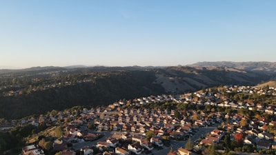 A suburban neighborhood with numerous houses spread across rolling hills. The landscape includes a mixture of residential areas and expansive green hills in the background, creating a contrast between urban and natural elements. The sky is clear with a light blue hue, adding to the peaceful setting.