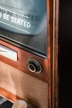 A vintage television set featuring a wooden exterior with a control dial and a retro speaker grill. There is a reflection on the glass showing text that partially reads 'TO BE SEATED.' The scene includes warm lighting and a lit candle nearby, adding a nostalgic ambiance.