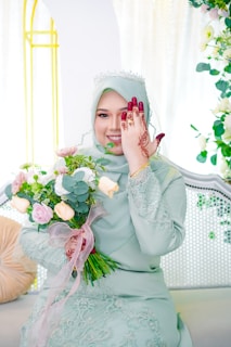 A woman in a light green dress with intricate detailing holds a bouquet of flowers. She is wearing a tiara and has henna designs on her hand, which she uses to cover one of her eyes. The background features light-colored decor with floral elements.