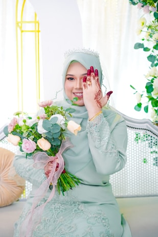 A woman in a light green dress with intricate detailing holds a bouquet of flowers. She is wearing a tiara and has henna designs on her hand, which she uses to cover one of her eyes. The background features light-colored decor with floral elements.