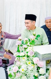 A group of people sitting together in what appears to be a celebratory or formal setting. One person is wearing a light green traditional outfit with a black cap, smiling warmly. A large bouquet of pink and white flowers is prominently displayed in the foreground, enhancing the festive atmosphere. The background consists of white drapes, contributing to the serene and joyful mood.